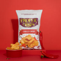 chilli crisps product image with red background chilli crips in a bowl on the side of the product and red chillis scattered on the side and infront of the product