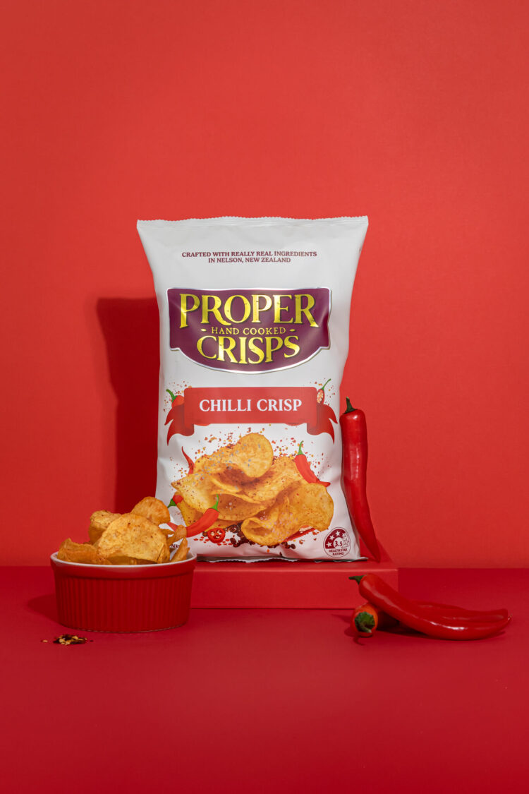 chilli crisps product image with red background chilli crips in a bowl on the side of the product and red chillis scattered on the side and infront of the product